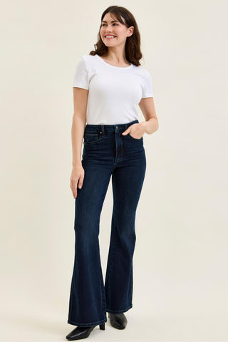 A woman modeling dark blue flared jeans with a white t-shirt and black shoes, standing against a plain beige studio background.