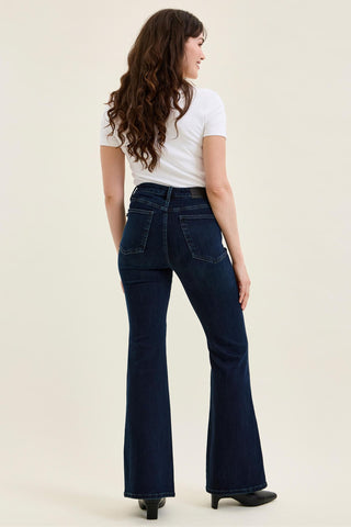 A woman shown from the back wearing dark blue high-waisted flare jeans, a white short-sleeve t-shirt, and black shoes against a plain light beige studio background.