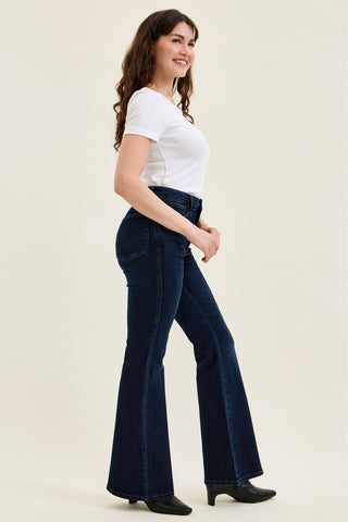 A woman modeling dark blue high-waisted flare jeans with a white t-shirt and black boots, standing against a solid light beige background.