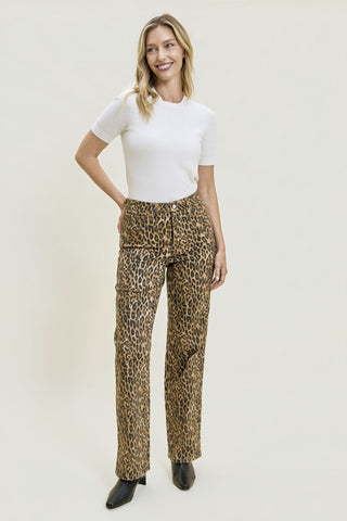 A woman modeling high-waisted leopard-print pants with a white short-sleeve top and black ankle boots, standing against a plain light beige studio background.