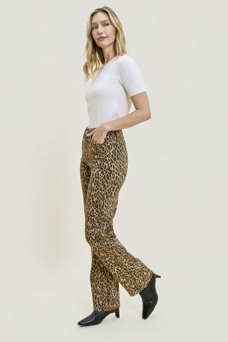 A woman modeling leopard-print flared pants with a white short-sleeve top and black pointed-toe heels, standing against a plain light beige studio background.