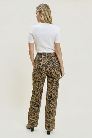 A woman shown from the back wearing leopard-print pants, a white short-sleeve top, and black heels against a plain light beige studio background.